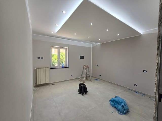 Stanza con pavimento grigio chiaro, controsoffitto bordato di luce fredda, faretti incassati, placche a parete e un termosifone sotto la finestra.