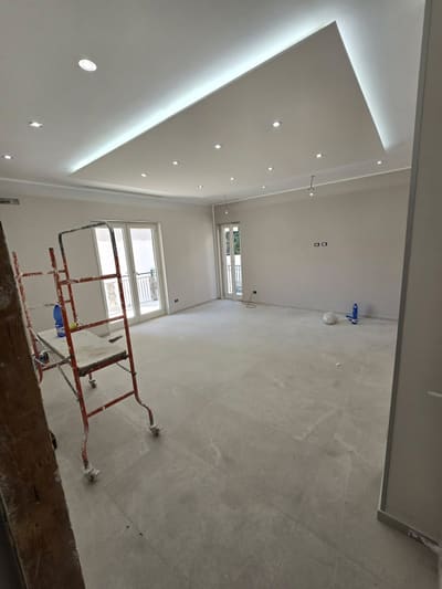 Grande ambiente con controsoffitto bordato di luce LED, faretti incassati, due porte finestre e un trabattello rosso a sinistra.