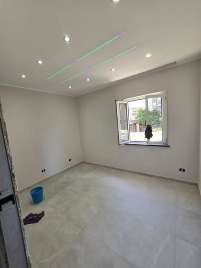 Stanza con faretti incassati e due sottili linee luminose parallele nel soffitto; finestra aperta sul fondo.