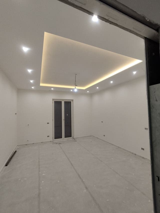 Stanza vuota con pareti bianche, controsoffitto con una cornice di luce calda al centro e faretti incassati; porta finestra sul fondo.