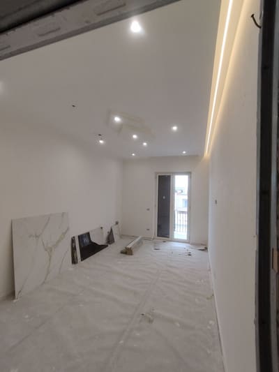 Stanza lunga e stretta con una linea di luce calda dove la parete destra incontra il soffitto, faretti e una porta finestra sul fondo.