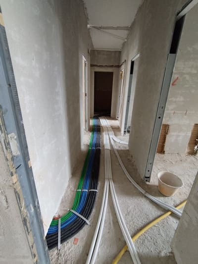 Corridoio grezzo con fasci di tubi flessibili blu, verdi e grigi stesi sul pavimento lungo il muro.