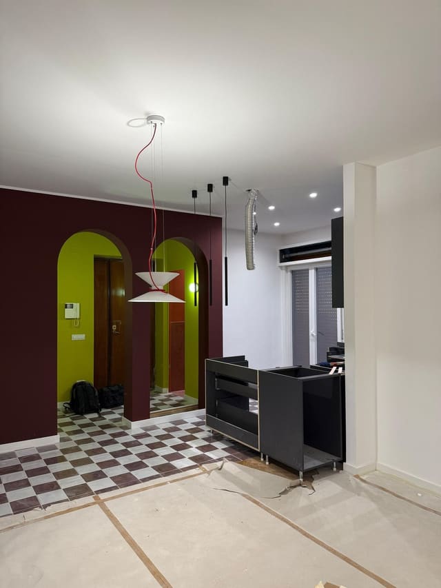 Ambiente con pareti bordeaux e verde acido, due archi, pavimento a scacchi, una lampada a sospensione con cavo rosso e sospensioni nere sottili.