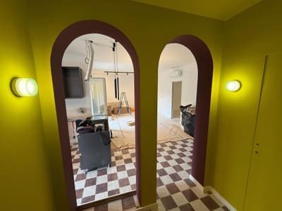 Parete gialla con due archi bordeaux e due applique tonde accese ai lati; oltre gli archi si vede la zona giorno in lavorazione.