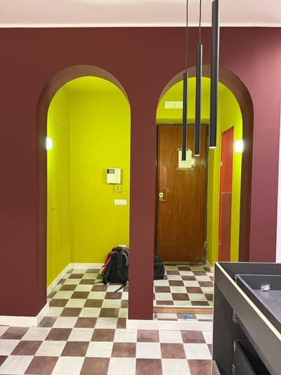 Ingresso con due archi, pareti bordeaux e verde acido, sospensioni nere sottili e un videocitofono accanto alla porta.