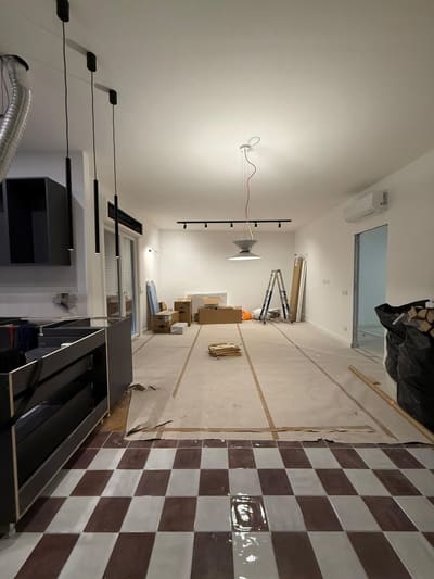 Ambiente lungo con sospensioni nere in primo piano, binario nero con faretti e lampada a sospensione sullo sfondo, pavimento a scacchi coperto in parte da teli.