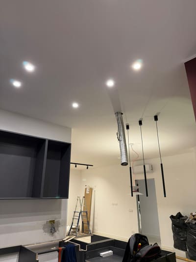 Zona giorno in lavorazione con faretti incassati nel soffitto, sospensioni nere sottili, un binario e un tubo flessibile in alluminio che scende dal soffitto.
