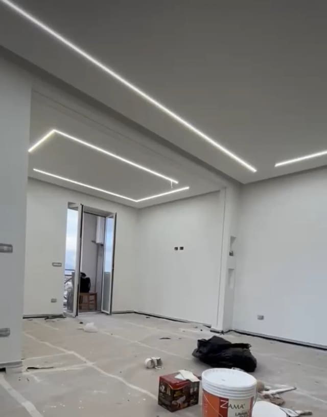 Stanza vuota in ristrutturazione con linee di luce LED incassate nel soffitto che formano rettangoli, pavimento coperto da teli e una porta finestra sul fondo.