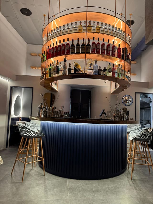 Espositore circolare per bottiglie sospeso e illuminato da luce arancione, sopra un bancone tondo scuro con una luce LED sotto il piano; sgabelli ai lati.