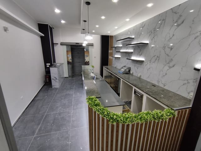 Bar finito con parete in lastre effetto marmo, mensole illuminate da strisce LED, faretti incassati nel controsoffitto, due lampade a sospensione e bancone con piante.