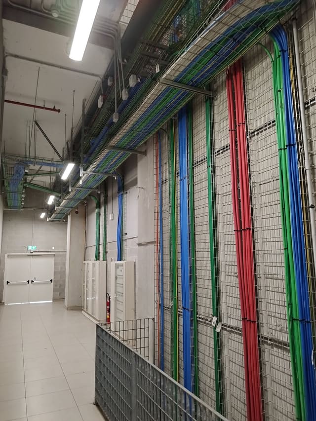 Corridoio tecnico con passerelle portacavi metalliche a soffitto e numerose tubazioni rosse, verdi e blu fissate alle pareti, illuminato da lampade lineari.