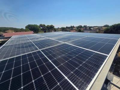 Primo piano di file di moduli fotovoltaici sotto un cielo azzurro, con tetti e alberi all'orizzonte.