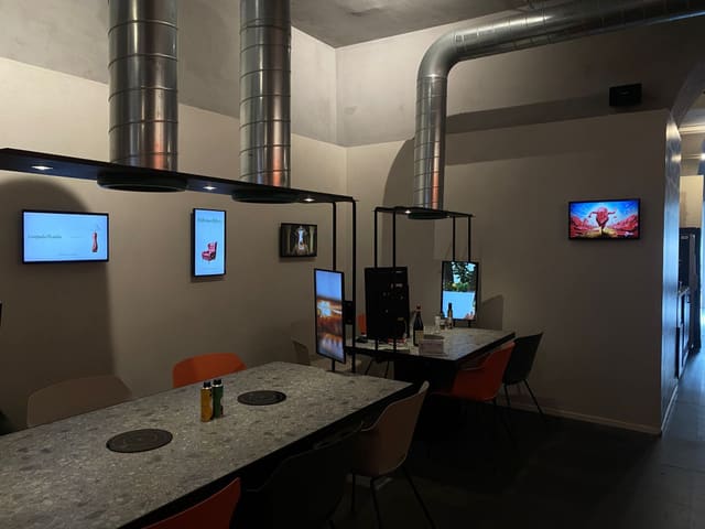 Sala di ristorazione con tavoli in pietra e sedie arancioni, strutture metalliche nere sopra i tavoli con faretti e schermi, altri schermi accesi alle pareti.