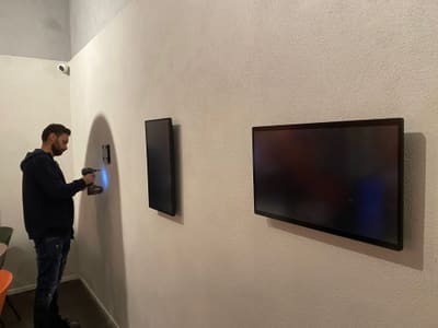 Un tecnico usa un trapano su una parete chiara, accanto a due schermi spenti montati a muro.