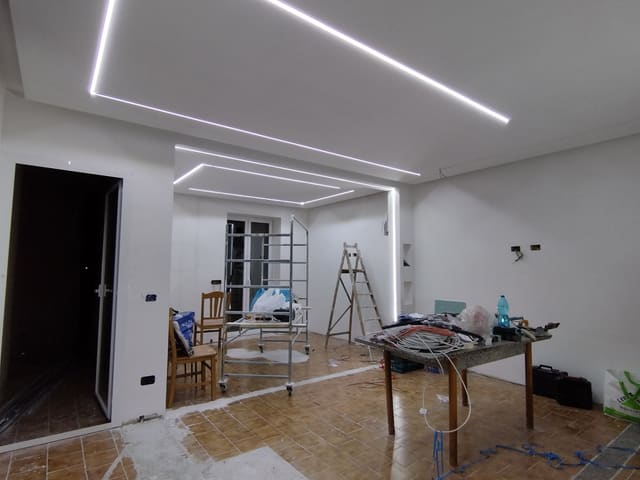 Stanze in ristrutturazione con linee LED incassate nel soffitto che formano due rettangoli, una linea verticale nell'angolo, un ponteggio mobile e un tavolo con cavi.