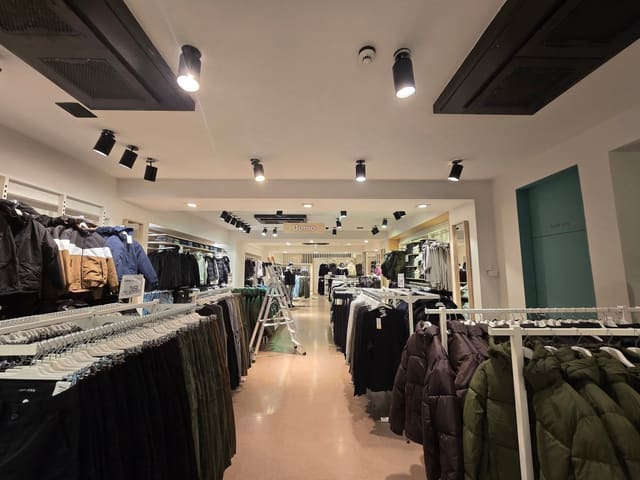 Corsia centrale di un negozio di abbigliamento con stender pieni di giacche ai lati e proiettori neri fissati al soffitto; sullo sfondo una scala.