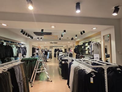 Reparto di un negozio di abbigliamento con proiettori neri cilindrici e faretti incassati nel soffitto bianco, sopra file di stender.