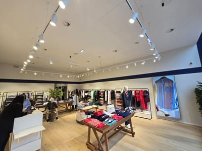 Negozio di abbigliamento con pavimento in legno, tavoli con capi piegati, manichini e binari bianchi sospesi con proiettori.