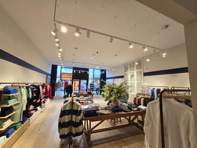 Sala di un negozio di abbigliamento vista verso la vetrina, con binari bianchi e proiettori sul soffitto, scaffali alle pareti e un tavolo centrale.