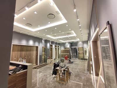 Vista laterale del negozio con uno specchio contornato di luce a destra, soffitto a gradoni illuminato, binario con faretti e cavalletti da lavoro.