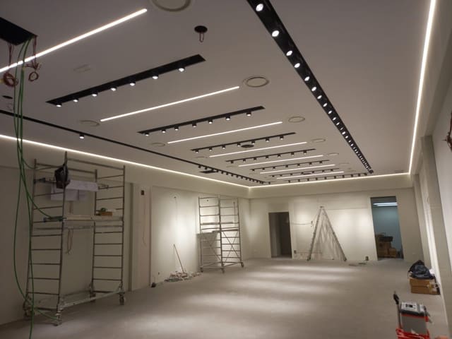 Soffitto di un negozio in allestimento con linee LED continue e file di binari neri con proiettori; sotto, ponteggi mobili.