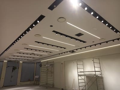 Soffitto con fasce nere di proiettori e linee LED parallele, gola luminosa lungo il perimetro e due ponteggi mobili sotto.