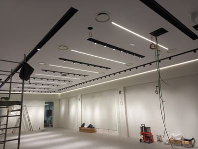 Soffitto con binari neri e linee LED accese; dal controsoffitto pendono cavi verdi, sotto ci sono una scala e un ponteggio.