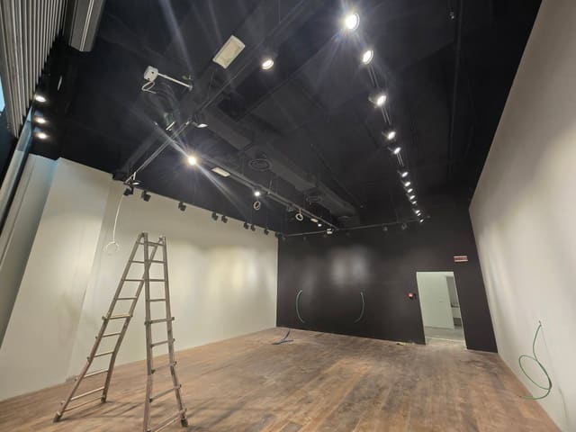 Locale vuoto con soffitto nero e impianti a vista, proiettori accesi su binari, parete di fondo nera, pavimento in legno e una scala in alluminio.
