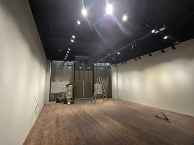 Locale commerciale con soffitto nero, file di proiettori su binario, pareti bianche e, sul fondo, la vetrina in allestimento con una scala.