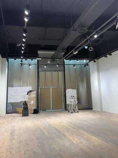 Locale con soffitto nero e impianti a vista, una fila di proiettori su binario a sinistra, vetrina sul fondo e materiali imballati vicino all'ingresso.
