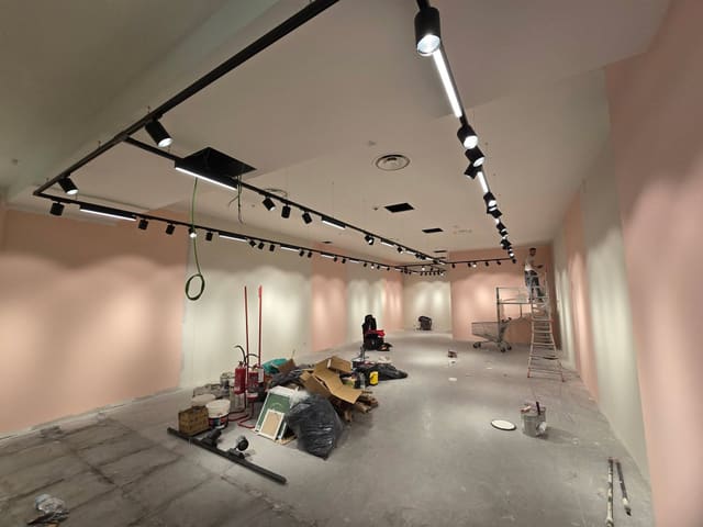 Locale commerciale vuoto con pareti rosa e bianche, binari neri sospesi con proiettori e linee luminose; a terra materiali da cantiere e un cavo verde che scende dal soffitto.