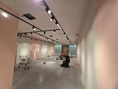 Locale lungo con pareti rosa, due file di binari neri con proiettori, una scala, un carrello e un tecnico al lavoro sul pavimento.