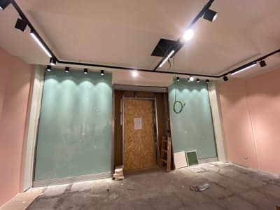 Parete di fondo con due pannelli verde acqua illuminati da proiettori su binario, porta d'ingresso chiusa da un pannello in legno, pareti rosa ai lati.