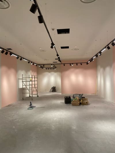 Locale vuoto con binari neri e proiettori lungo il perimetro del soffitto, pareti rosa illuminate e un ponteggio mobile al centro.