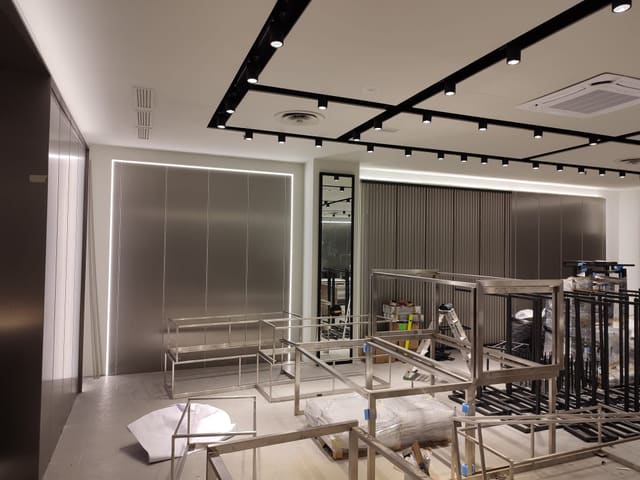Negozio in allestimento con soffitto bianco diviso da binari neri pieni di proiettori, un condizionatore a cassetta e una parete metallica contornata da una linea luminosa.