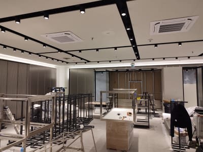 Soffitto con griglia di binari neri e due condizionatori a cassetta; in basso, espositori metallici e scatoloni.