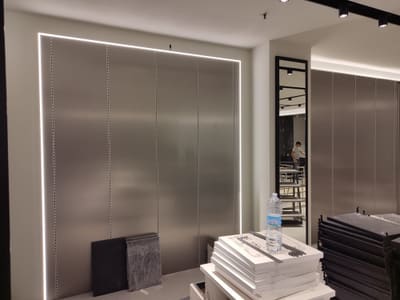 Parete di pannelli metallici grigi contornata da una sottile linea luminosa; a destra uno specchio con cornice nera, a terra mobili imballati.