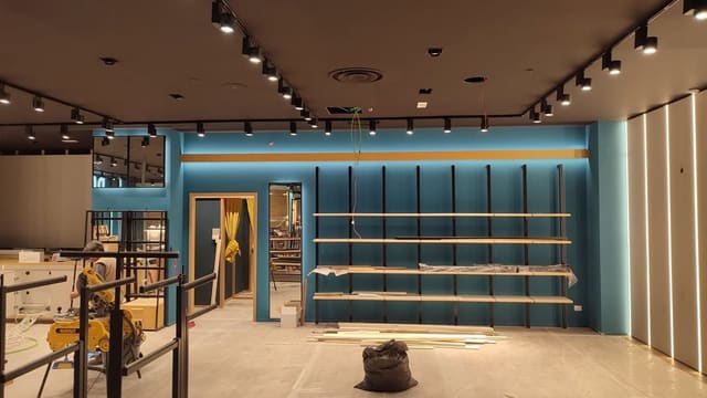 Negozio in allestimento con soffitto scuro e binari neri con proiettori lungo i bordi, parete blu con mensole illuminata dall'alto e, a destra, pareti chiare con linee luminose verticali.