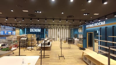 Ampio negozio con pavimento in legno, soffitto scuro con file di binari e proiettori, pareti blu e chiare con linee luminose verticali; espositori e scatoloni al centro.