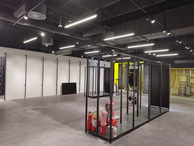 Ampio locale commerciale con soffitto nero a vista e condotte d'aria, file di lampade lineari LED sospese, binari con faretti, pareti bianche e strutture espositive metalliche in montaggio.