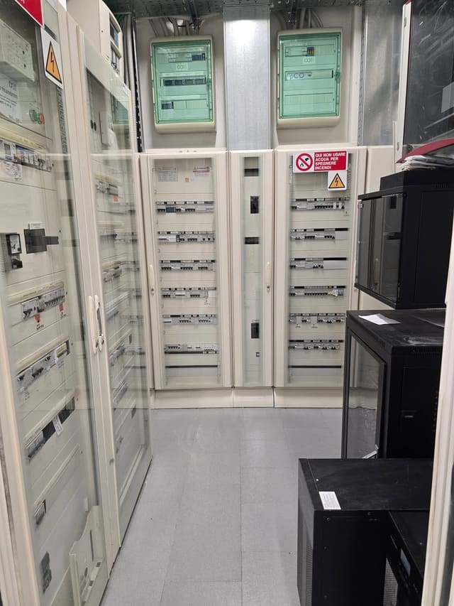 Locale tecnico con diversi quadri elettrici affiancati, pieni di interruttori modulari, cartelli di sicurezza e armadi neri a destra.