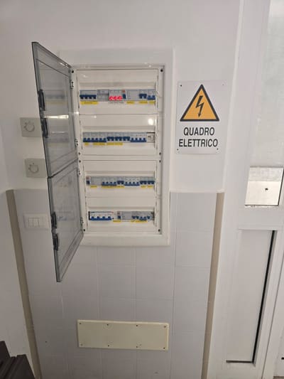 Quadro elettrico incassato in una parete piastrellata, con lo sportello trasparente aperto su file di interruttori e un cartello giallo con la scritta "quadro elettrico".