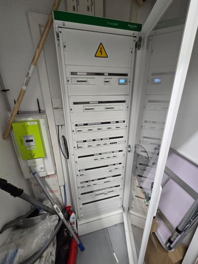 Armadio elettrico alto e bianco con la porta aperta e un cartello di pericolo in alto; accanto, un contenitore giallo-verde a parete.