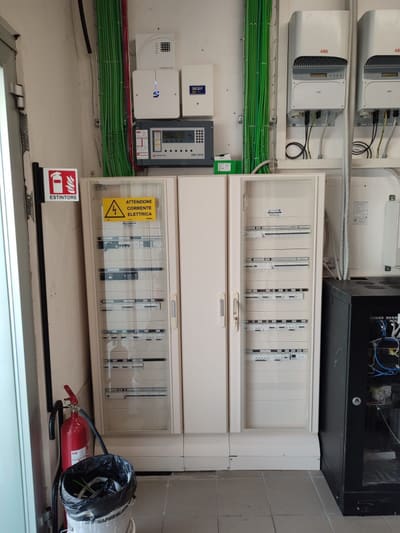 Quadri elettrici a due ante con cartello di avvertenza, un armadio rack nero, tubi verdi per i cavi lungo la parete e un estintore.
