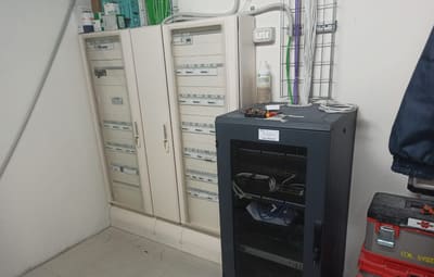 Quadri elettrici bianchi a parete e un armadio rack nero con cavi di rete, accanto a una cassetta degli attrezzi rossa.