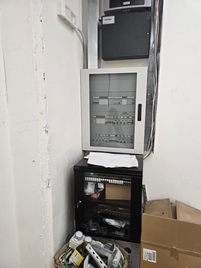 Quadro elettrico grigio a parete con sotto un piccolo armadio rack nero aperto pieno di cavi; scatoloni a terra.