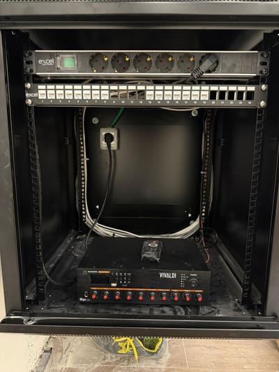 Interno di un armadio rack nero con una multipresa, un pannello di connessione per i cavi di rete e un apparecchio con manopole in basso.