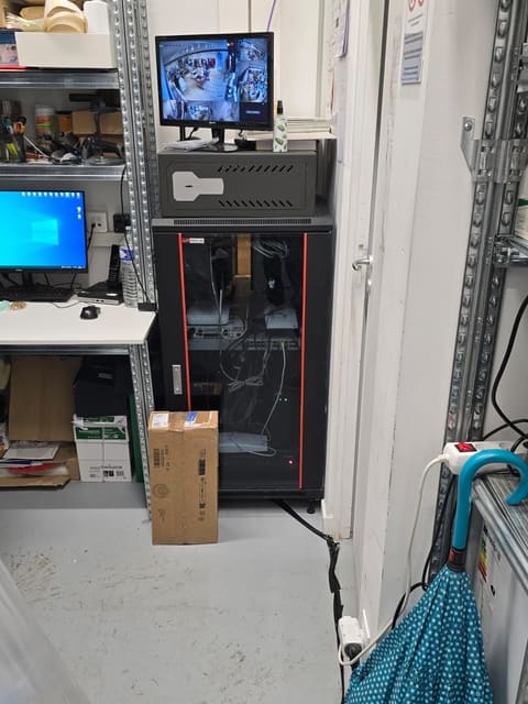 Monitor che mostra le immagini di più telecamere, appoggiato sopra un armadio rack nero, accanto a una scrivania con un computer.