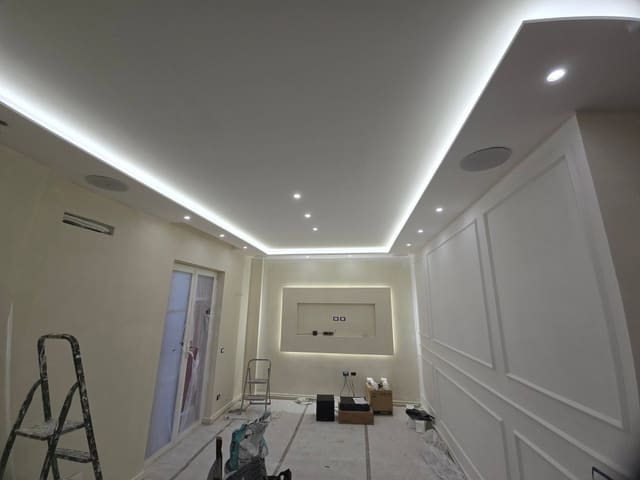 Stanza in ristrutturazione con controsoffitto illuminato lungo il bordo da una striscia LED, faretti, pareti con cornici decorative e una nicchia illuminata sul fondo.