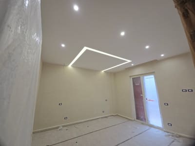 Stanza con faretti incassati e una linea luminosa ad angolo nel soffitto; porta finestra sul fondo e teli protettivi a terra.